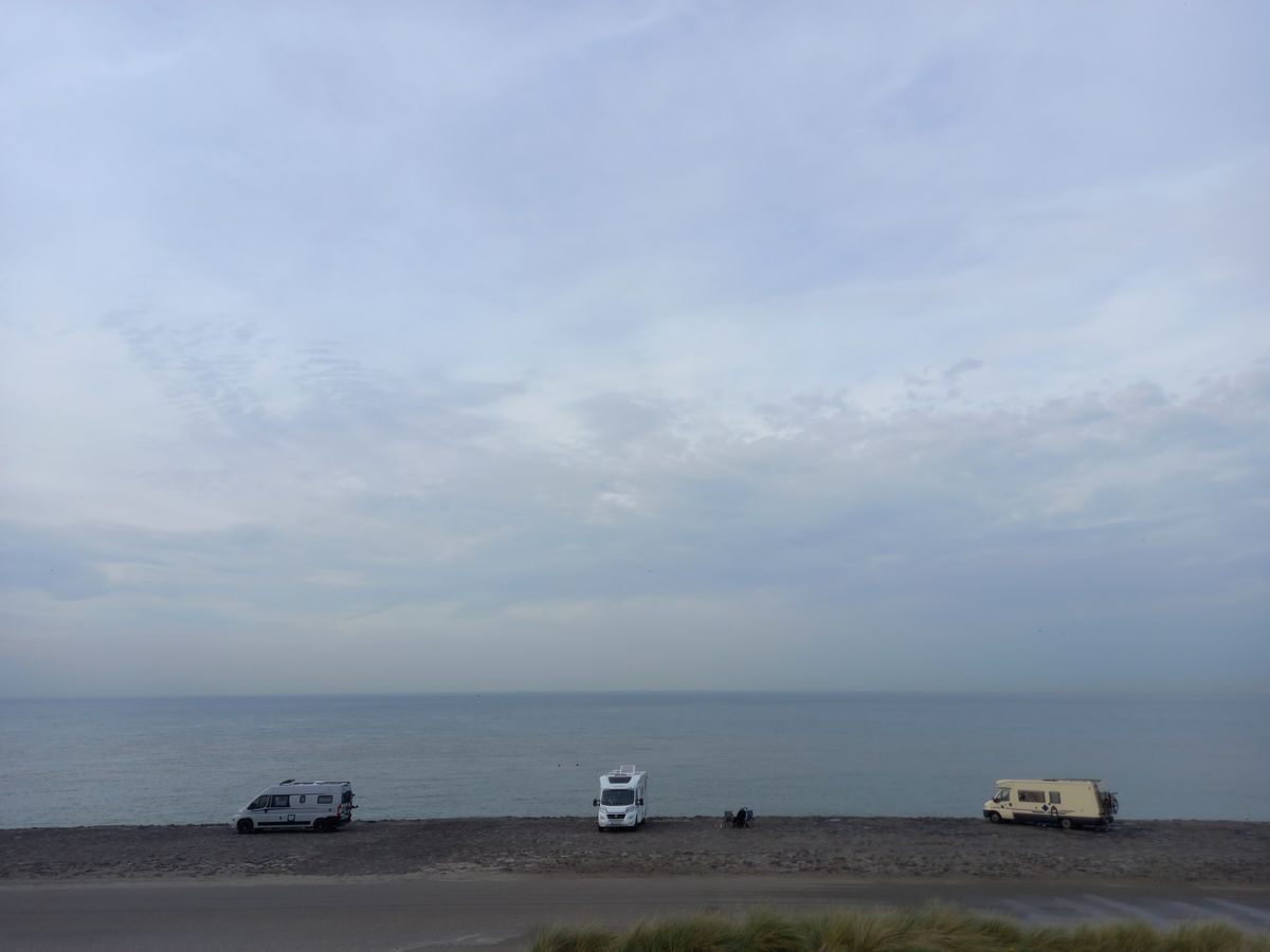 Drei Wohnmobile stehen nebeneinander auf einem Kiesstreifen direkt am ruhigen Meer, darüber ein weiter, diesiger Himmel